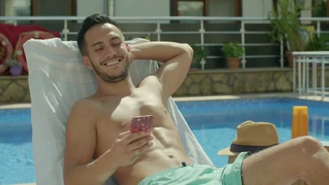 A brunette man spends time with his phone while sunbathing by the pool. Stock Footage 144315233