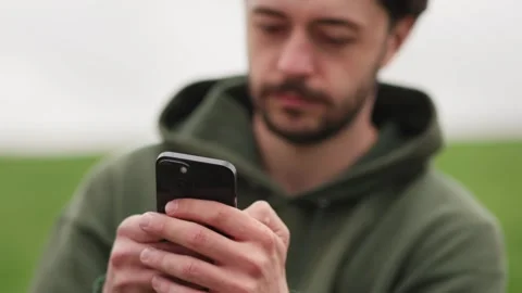 A brunette man uses a phone while sitting in a park. A bearded man in a green Stock Footage 240726467