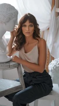 Brunette model in elegant attire posing by a Grecian bust under soft daylight 库存照片