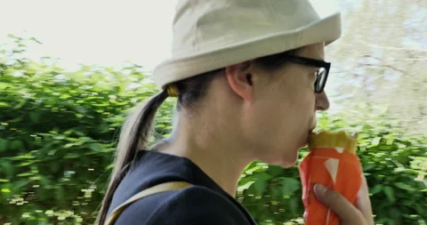 Brunette with ponytail and glasses eats fast food on  go. A woman with a hat Stock Footage 276614372