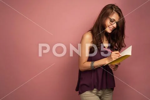 A brunette stands on a pink background in a purple top and glasses, smiling Stock Photos