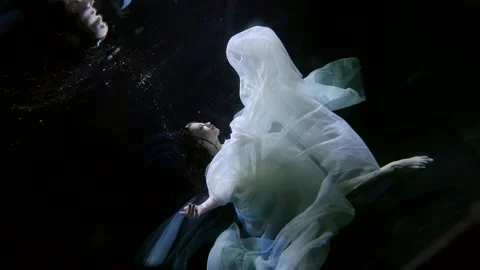 A brunette in a white translucent fabric floated under the surface of the dark Stock-Footage 205031898