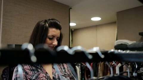 Brunette woman in checkered shirt comes to rack with hangers in store Stock Footage