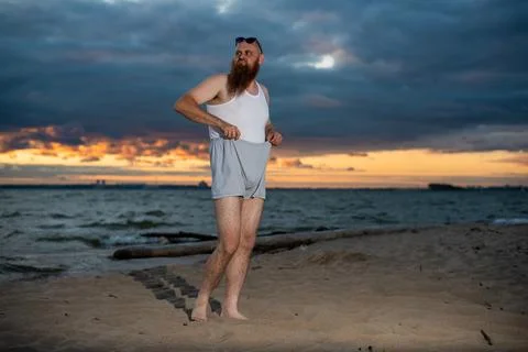 A brutal bald man posing on the beach at sunset parody glamorous chick Stock Photos