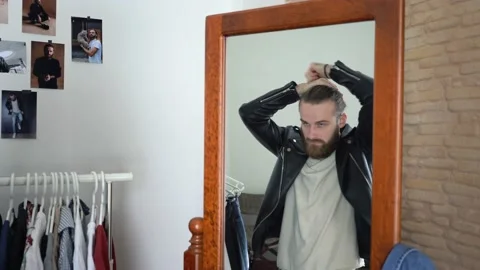 Brutal beard man in a Leather Jacket  is looking in a mirror tying his hair Stock Footage 147893677