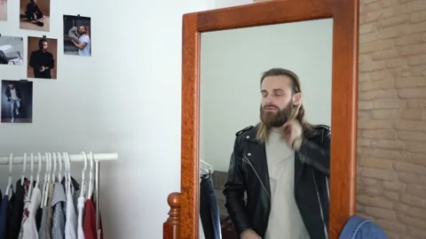 Brutal beard man in a Leather Jacket  is looking in a mirror Untwisting his hair Stock Footage 147893778