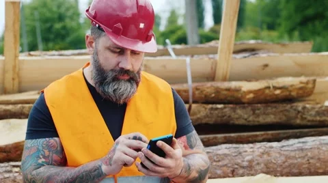 Brutal builder with a beard and tattoos sitting on the logs, using a mobile Stock Footage 62940636