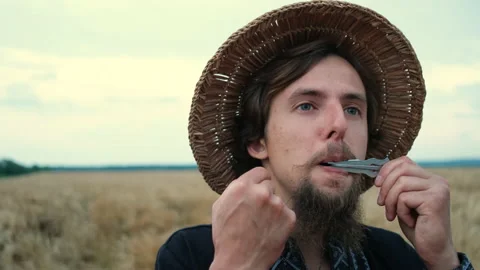 A brutal man with a beard and a straw hat in wheat plays an ancient instrument Stock Footage 136823726
