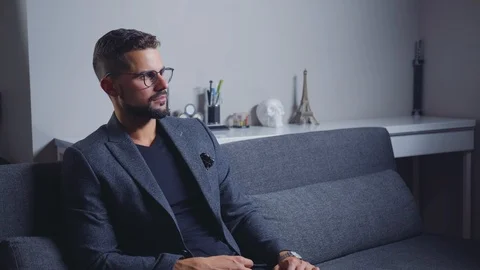 Brutal man with a beard sits on a sofa and looks at the nachas time Stock Footage 118157690