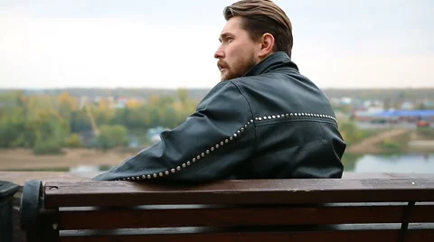 Brutal man in a leather jacket sitting on a park bench Stock Footage 68622838