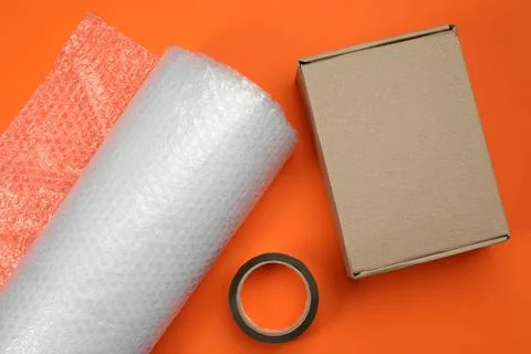 Bubble wrap roll, cardboard box and tape on orange background, flat lay Stock Photos