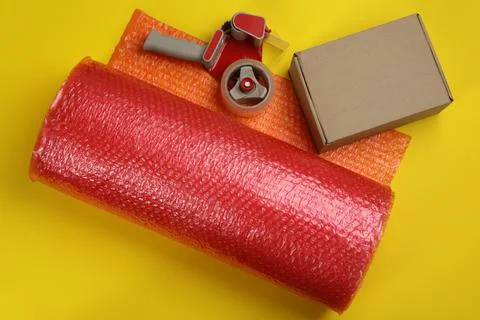 Bubble wrap roll, cardboard box and tape dispenser on yellow background, flat Stock Photos