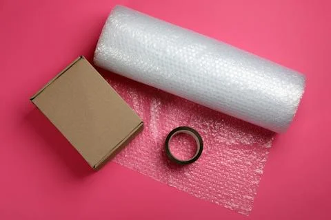Bubble wrap roll, cardboard box and tape on pink background, flat lay Stock Photos