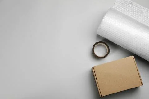 Bubble wrap roll, cardboard box and tape on light background, flat lay. Space Stock Photos