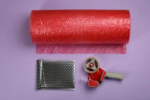 Bubble wrap roll, padded envelope and tape dispenser on lilac background, fla Stock Photos