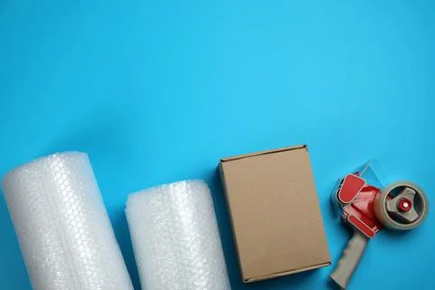 Bubble wrap rolls, cardboard box and tape dispenser on light blue background, Stock Photos