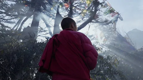 Buddhist monk in red robe takes off yellow hat. Sun shine in dense smoking mist Stock Footage 167418568