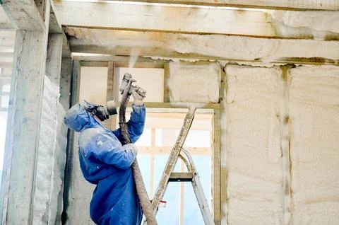 Builder applying spray foam insulation to overhead wall section in unfinished Фото