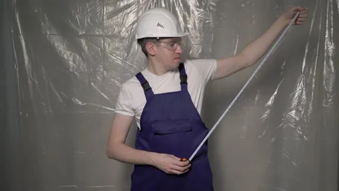 Builder in blue overalls and white hard hat helmet and glasses with tape-measure Vidéo 175998186