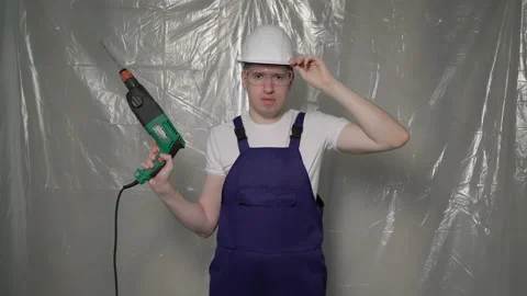 Builder in blue overalls and white hard hat helmet and glasses with perforator. Vidéo 176018319