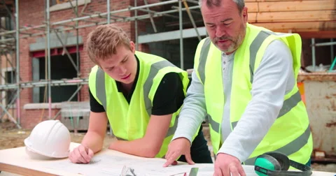 Builder On Building Site Discussing Work With Apprentice Vídeos de archivo 56221222