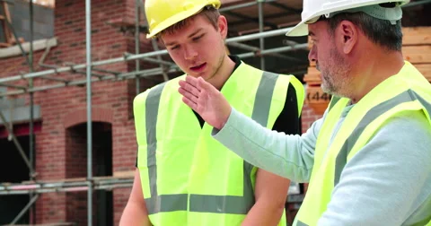 Builder On Building Site Discussing Work With Apprentice Vídeos de archivo 56221320