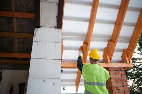 Builder fits insulation layer between timber roof joists, modern house cons.. Stock Photos