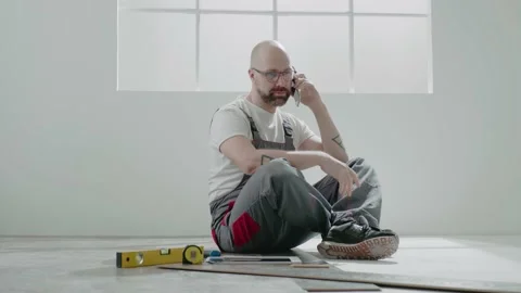 Builder with glasses calls on mobile phone sitting on floor Stock Footage 149892248