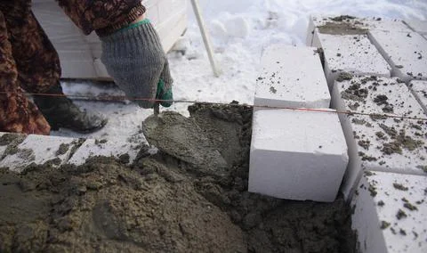 A builder in mittens puts gray bricks in cement in straight rows. 스톡 사진