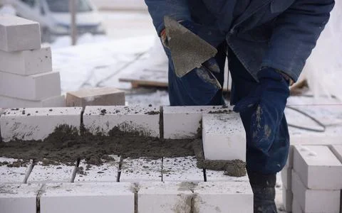 A builder in mittens puts gray bricks in cement in straight rows. Stock Photos
