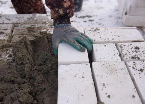 A builder in mittens puts gray bricks in cement in straight rows. Stock Photos