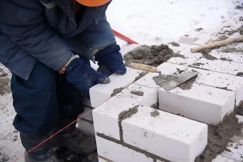 A builder in mittens puts gray bricks in cement in straight rows. Stock Photos