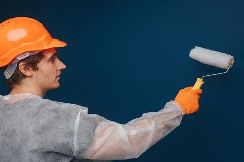 Builder is using roller to paint wall, isolated over blue background Stock Photos