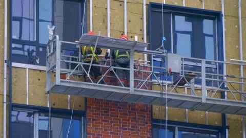 A builders in a cradle on cables fastens decorative cladding panels on the wall  Stock Footage 229822834