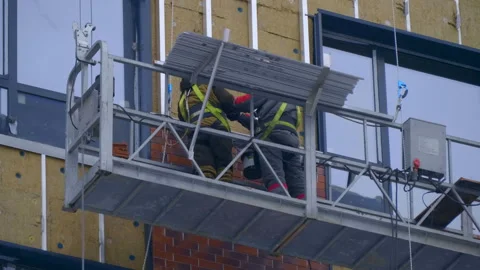 A builders in a cradle on cables fastens decorative cladding panels on the wall  Stock Footage 229824784