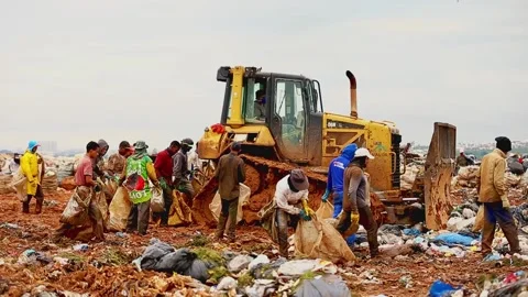 Bulldozer plows piles of garbage at a dump, poor people follow the 스톡 동영상 136839425
