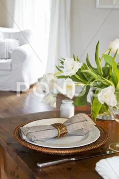 A Bunch Of White Tulips In A Glass Vase; A Place Setting With A Basket Plate To