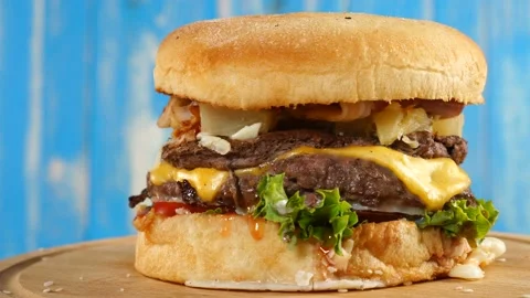 Burger, with a beef, cheese, and vegetables rotates on a wooden Board. On blue Stock Footage 112044592