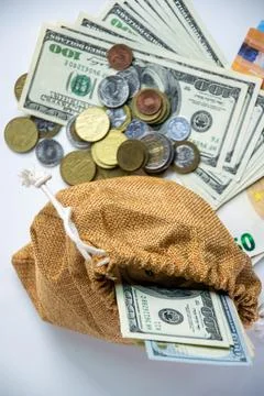 A burlap bag sits on a white surface with US dollar bills and coins spillin.. Stock Photos