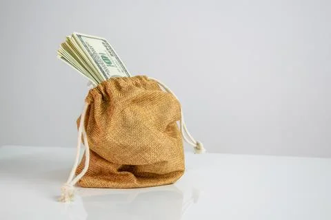 A burlap sack with a drawstring sits on a white surface with a stack of US .. Stock Photos
