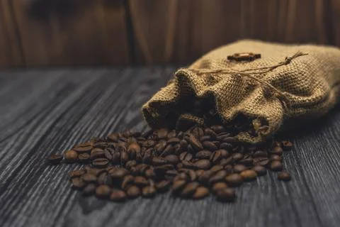 Burlap sack spilling coffee beans on dark wooden surface Stock Photos