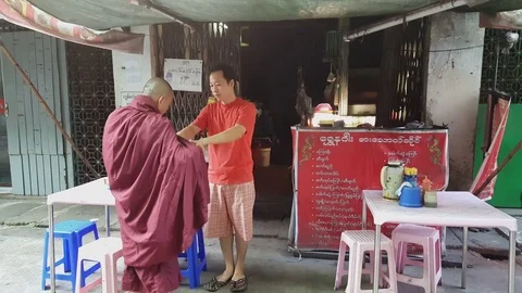 Burmese man gives alms to robed monk at coffee shop, tables, Yangon, Myanmar Stock Footage