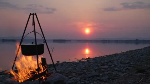 A burning campfire with a hanging pot on a tripod. Dramatic sunset on the lake. Stock Footage 242285140