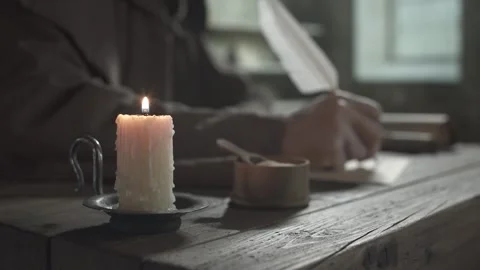 Burning candle on a rustic desk against a blurred background of a writing monk Stock Footage 339631025