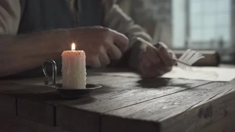Burning wax candle on a table with a scholar sharpening a quill pen in backgroun Vidéo 342016575