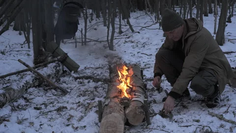 Bushcraft man maintaining long fire log in winter forest survival camp Stock Footage 334373362