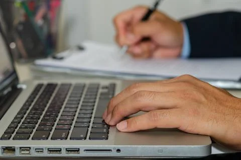 Business man hand typing a computer and holding a pen to write business doc.. Stock Photos