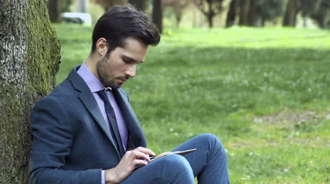 Business man sit under a tree in a city park typing on his tablet Video stock 64727993