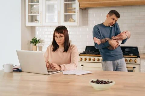 Business mom working in the kitchen while young father carrying their baby Stock Photos