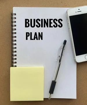 Business plan text on note pad with smart phone, pen and sticky note background. Stock Photos